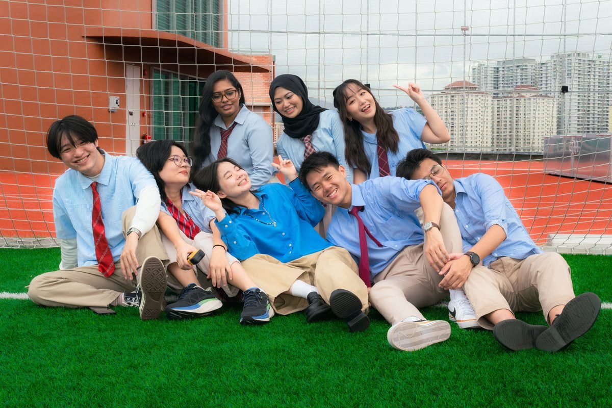 A group of friends in blue shirts and red ties pile up on rooftop grass in front of a goal net, laughing.