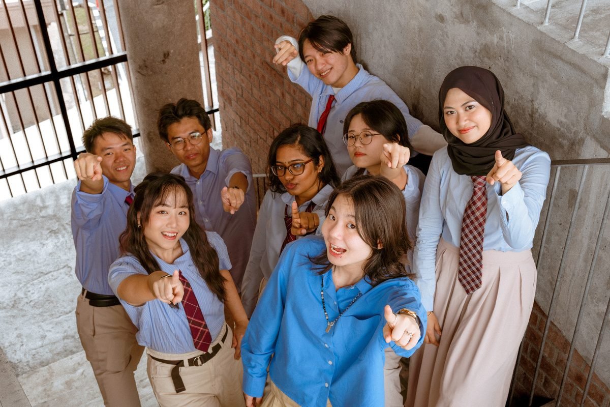 A group of friends in blue shirts and red ties on a concrete staircase, all pointing at the camera from above.