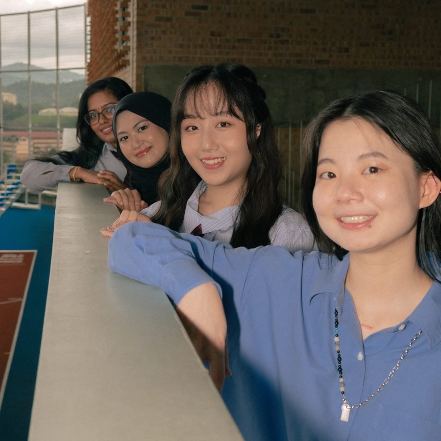Four friends lean on a rooftop railing and smile at the camera, with a red running track behind them.