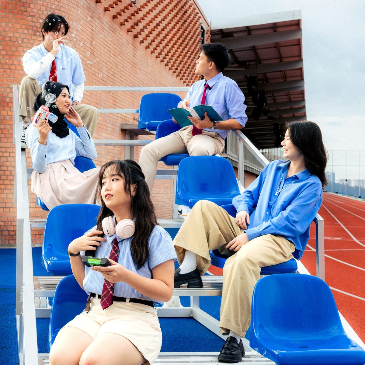 Friends lounge on blue stadium seats, one with pink headphones and a camera, another reading a book.