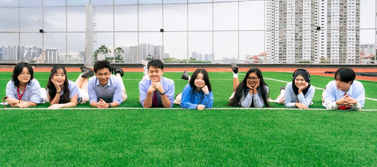 A row of friends lie on their stomachs on the grass, chins on their hands, looking at the camera.