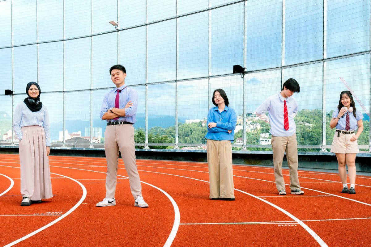 Five friends stand spread out on a red running track in front of a glass wall and city skyline.