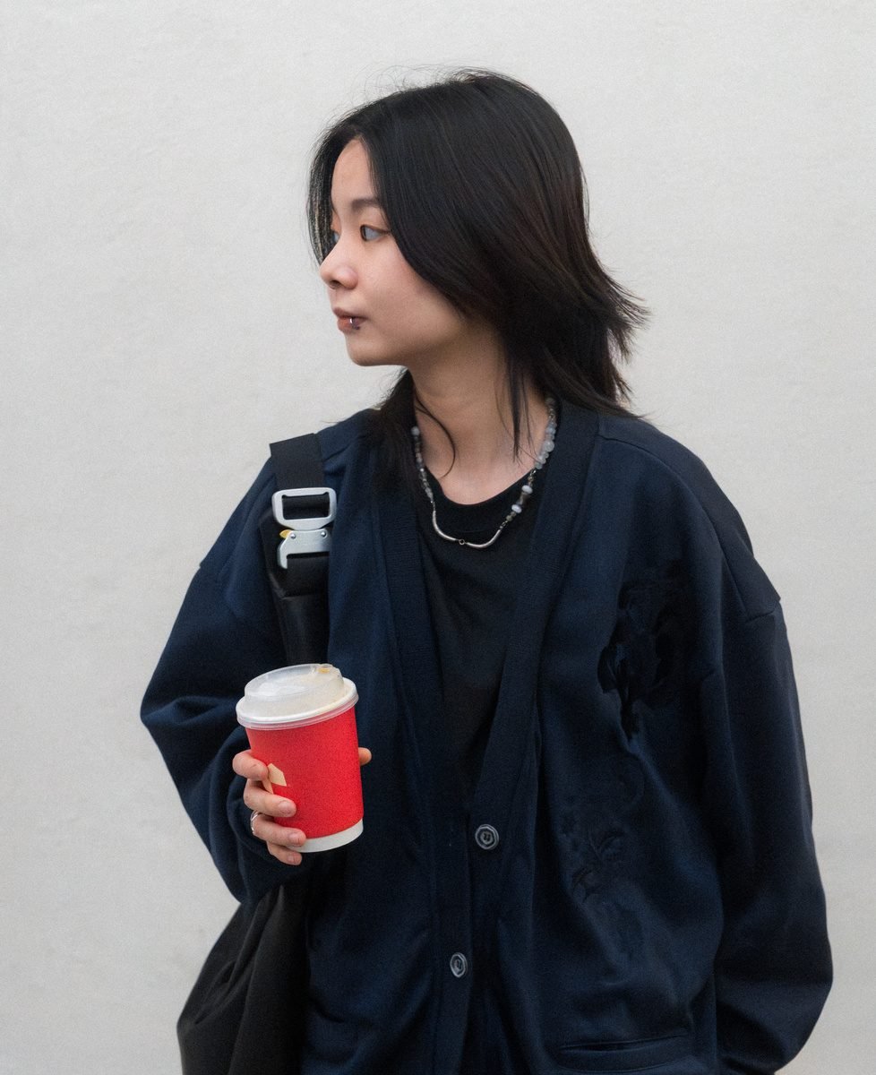 A woman in a navy cardigan looks away from the camera against a white wall, holding a red coffee cup.
