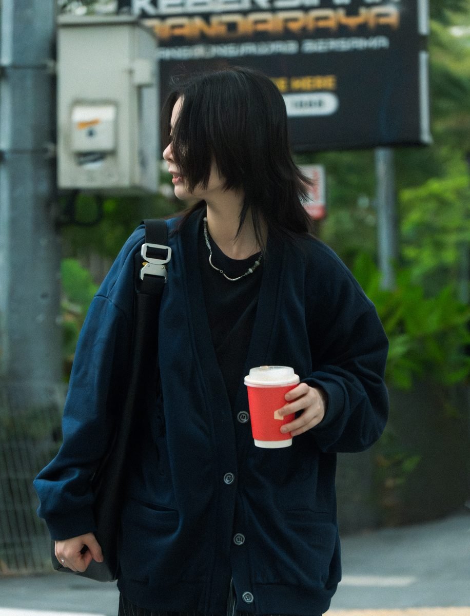 The woman crosses a street, glancing off to the side with the red cup in hand and green trees behind her.