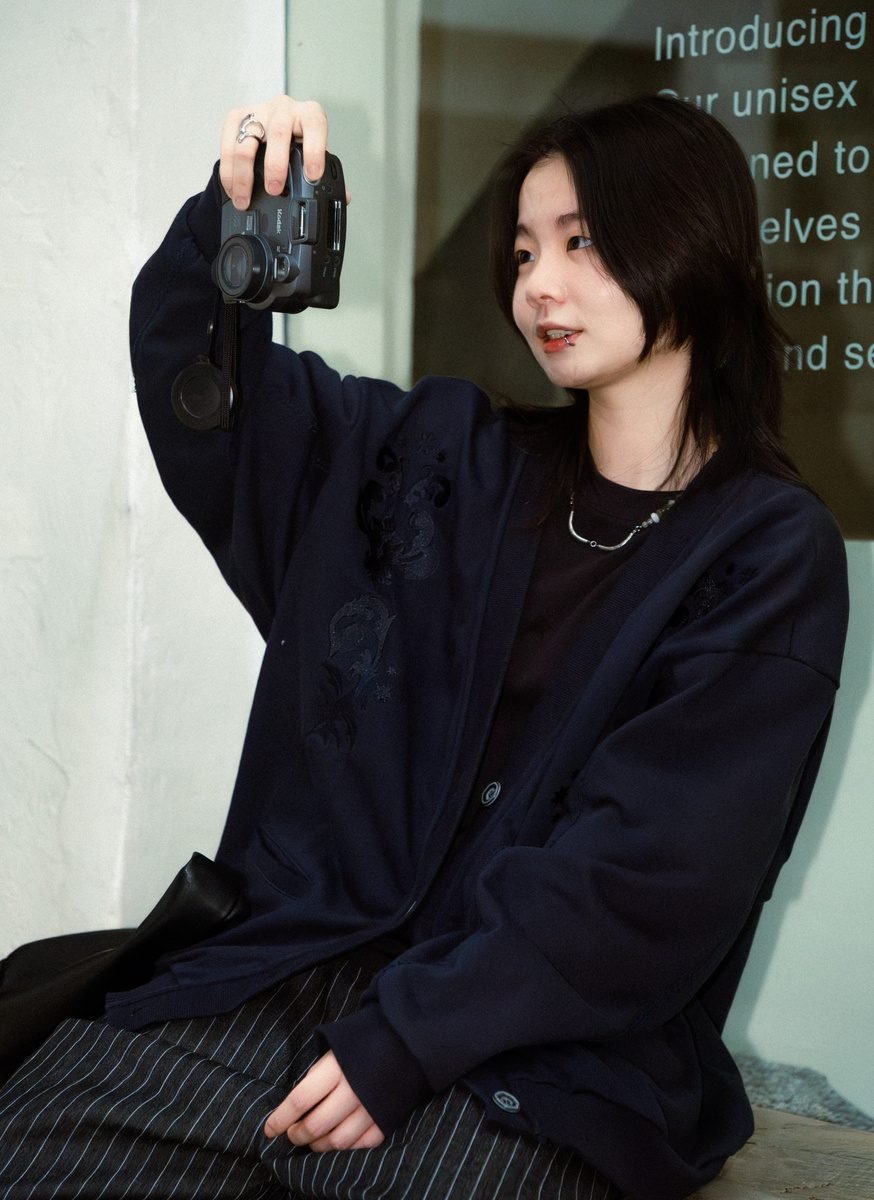 Seated, she holds up a compact film camera to take a picture, with shop window text reflected behind her.