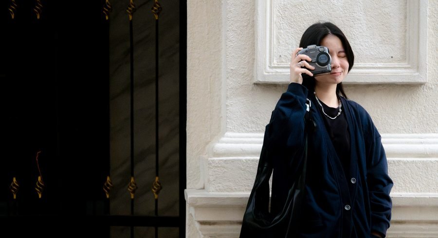 She covers one eye with a film camera, leaning on a cream wall beside an ornate black gate.