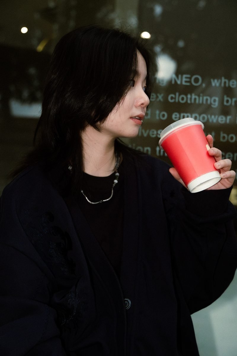 Side profile of her sipping from the red cup in front of a dark shop window.