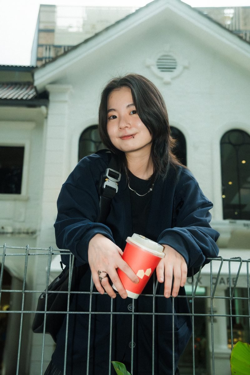She leans on a wire fence in front of a white colonial-style building, smiling, with the cup tilted in her hand.