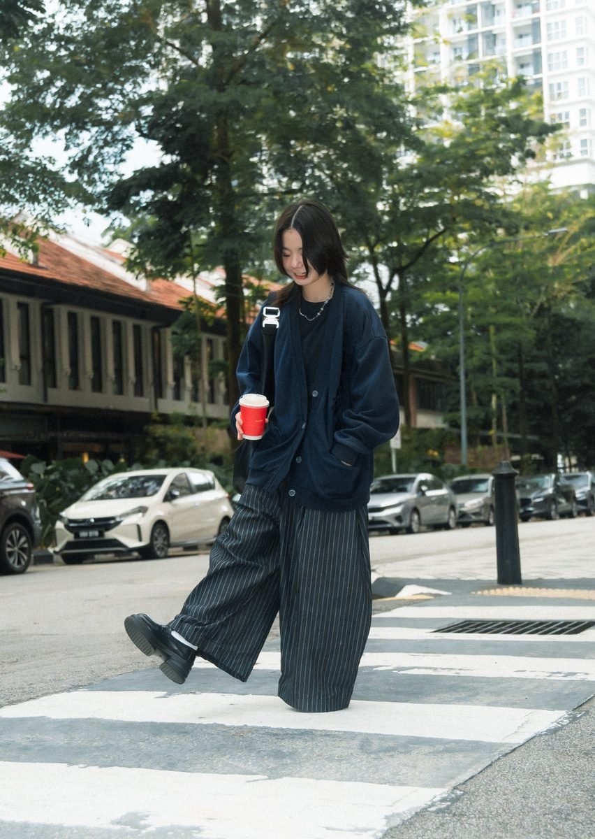 Mid-stride on a zebra crossing in wide pinstripe trousers, with shophouses and parked cars behind.