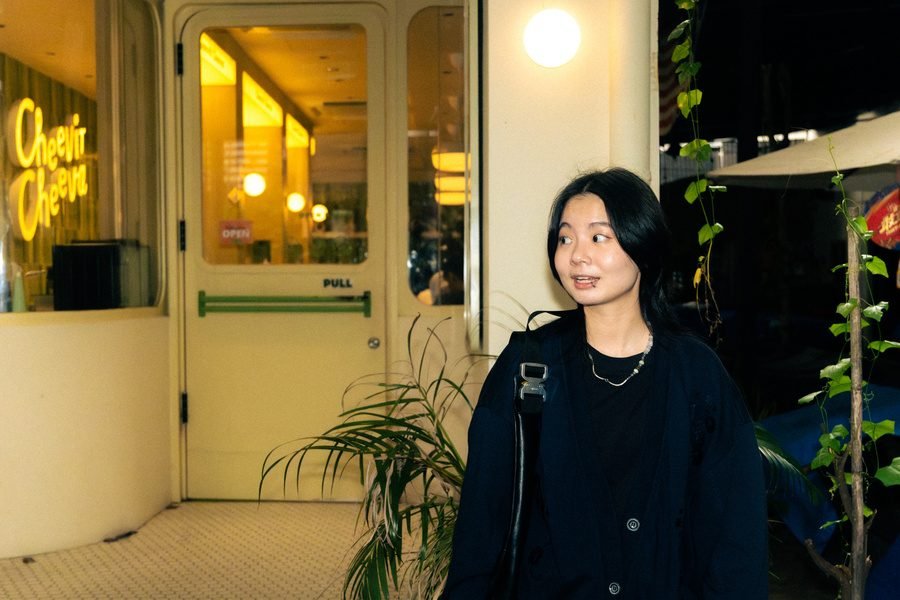 She stands outside a warmly lit cafe at night, looking off to one side beside a palm.