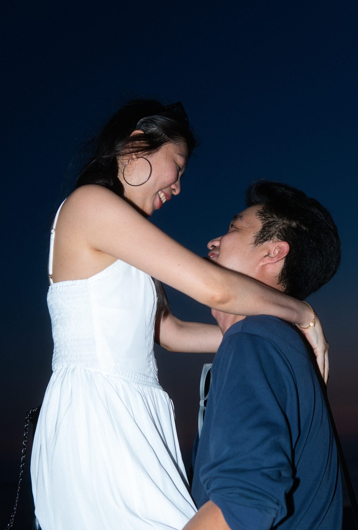 A woman in a white dress laughs as she looks down at a man in a navy shirt, lit by flash against a deep blue evening sky.