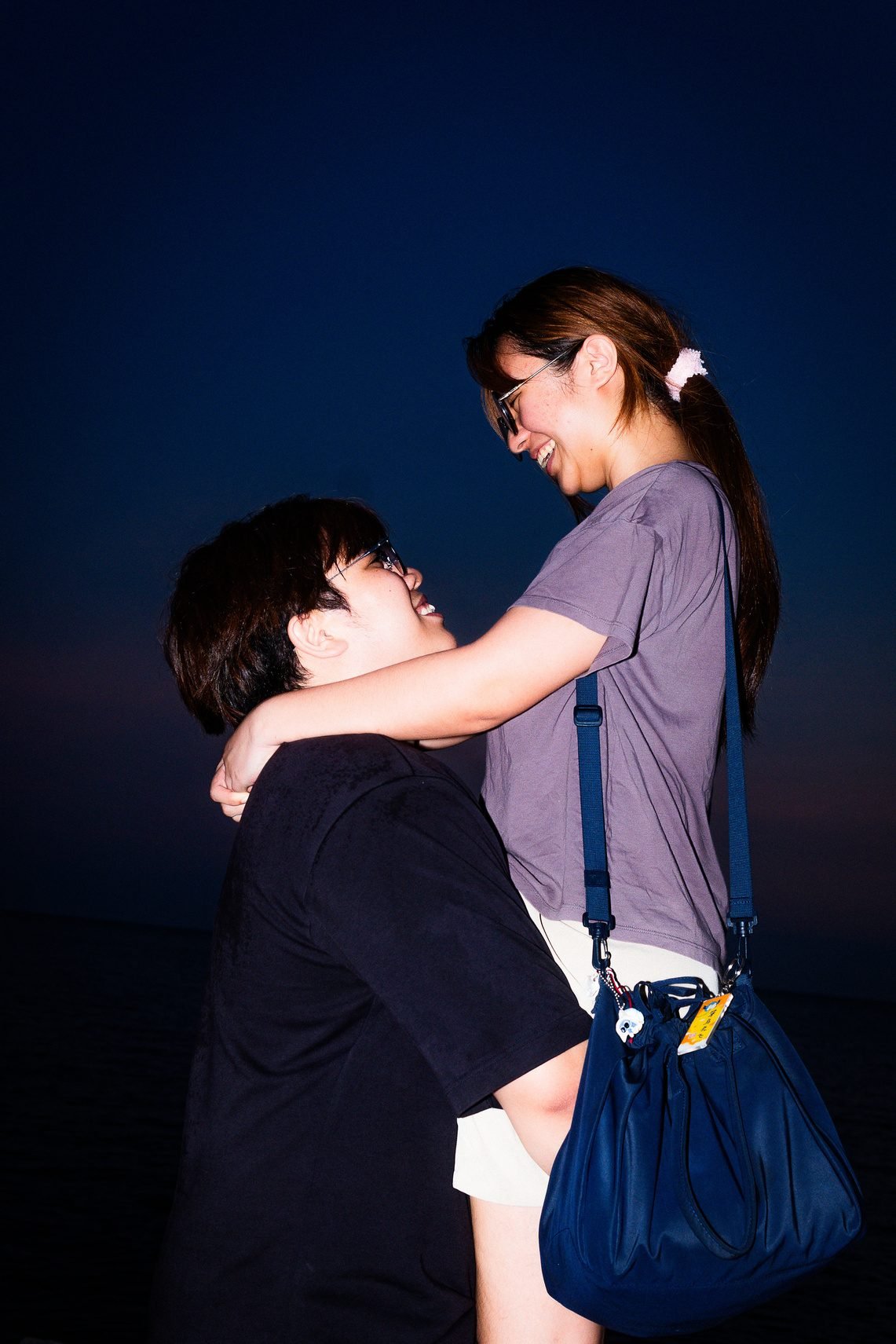 A man looks up and laughs as a woman in a grey tee with a navy bag wraps her arm around his neck, lit by flash against a dark blue sky.