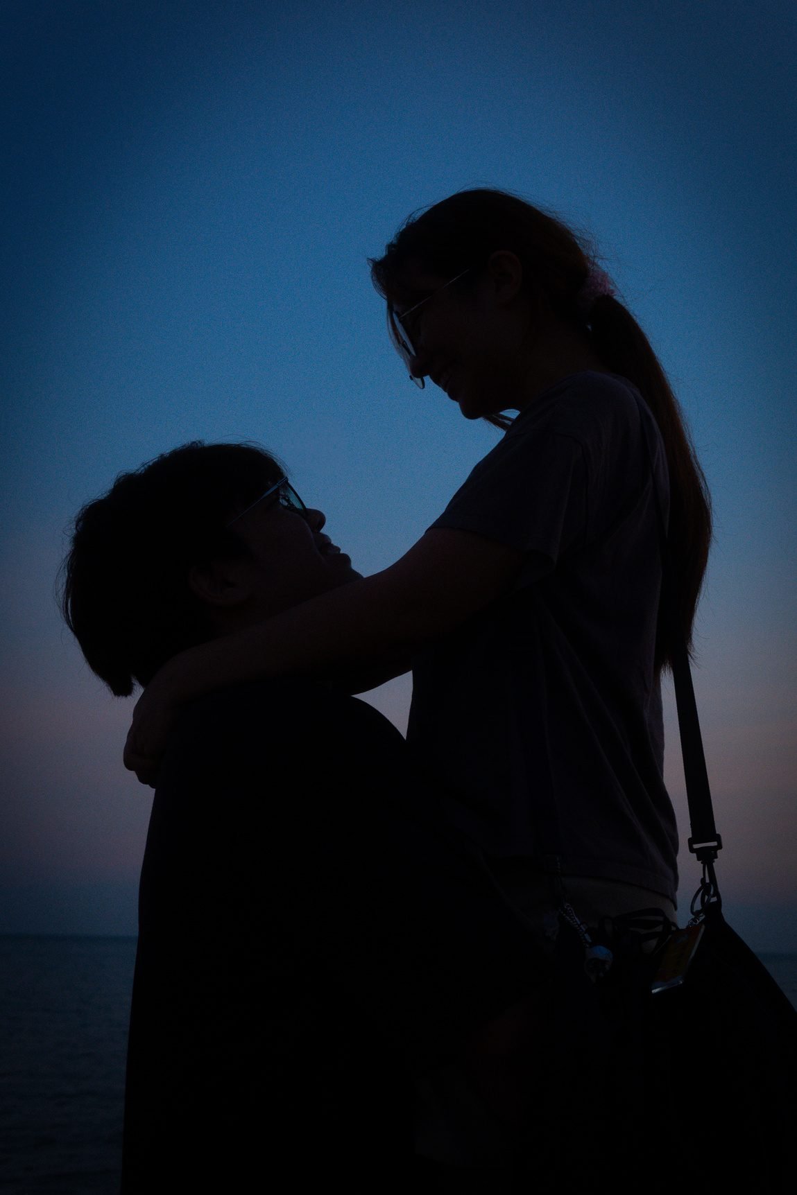The same couple as silhouettes, with the sea and a faint pink horizon behind them.