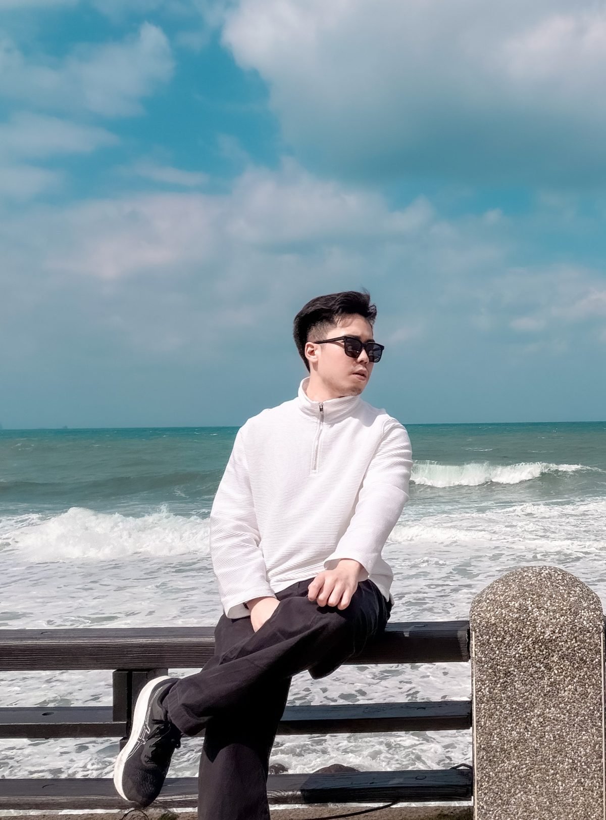Wing Keen in sunglasses and a white zip-neck sweater, sitting on a wooden railing by a rough blue-green sea under a cloudy sky.
