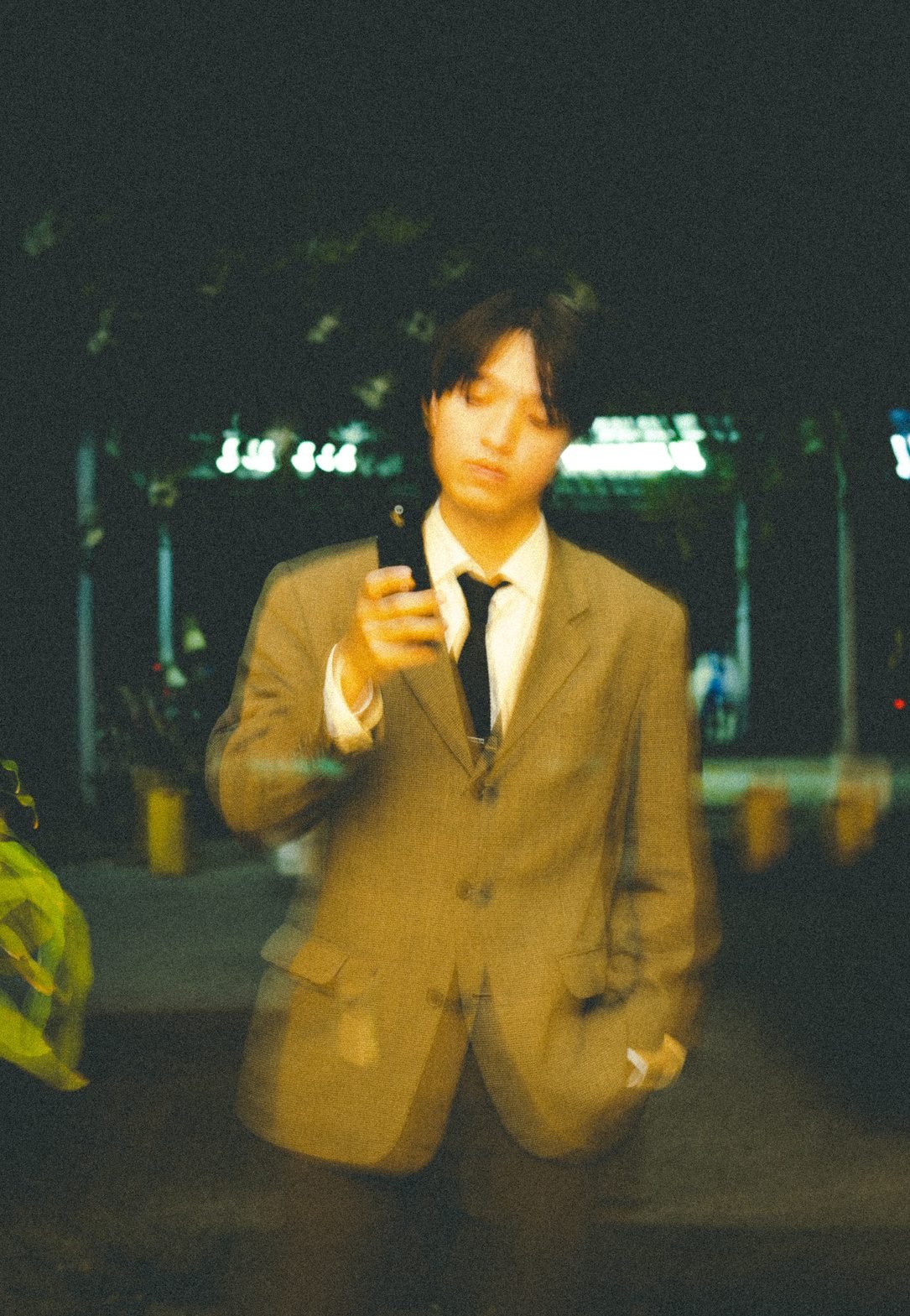 A man in a tan suit holds up his phone and looks down at it, out of focus and glowing yellow-green against a dark street.