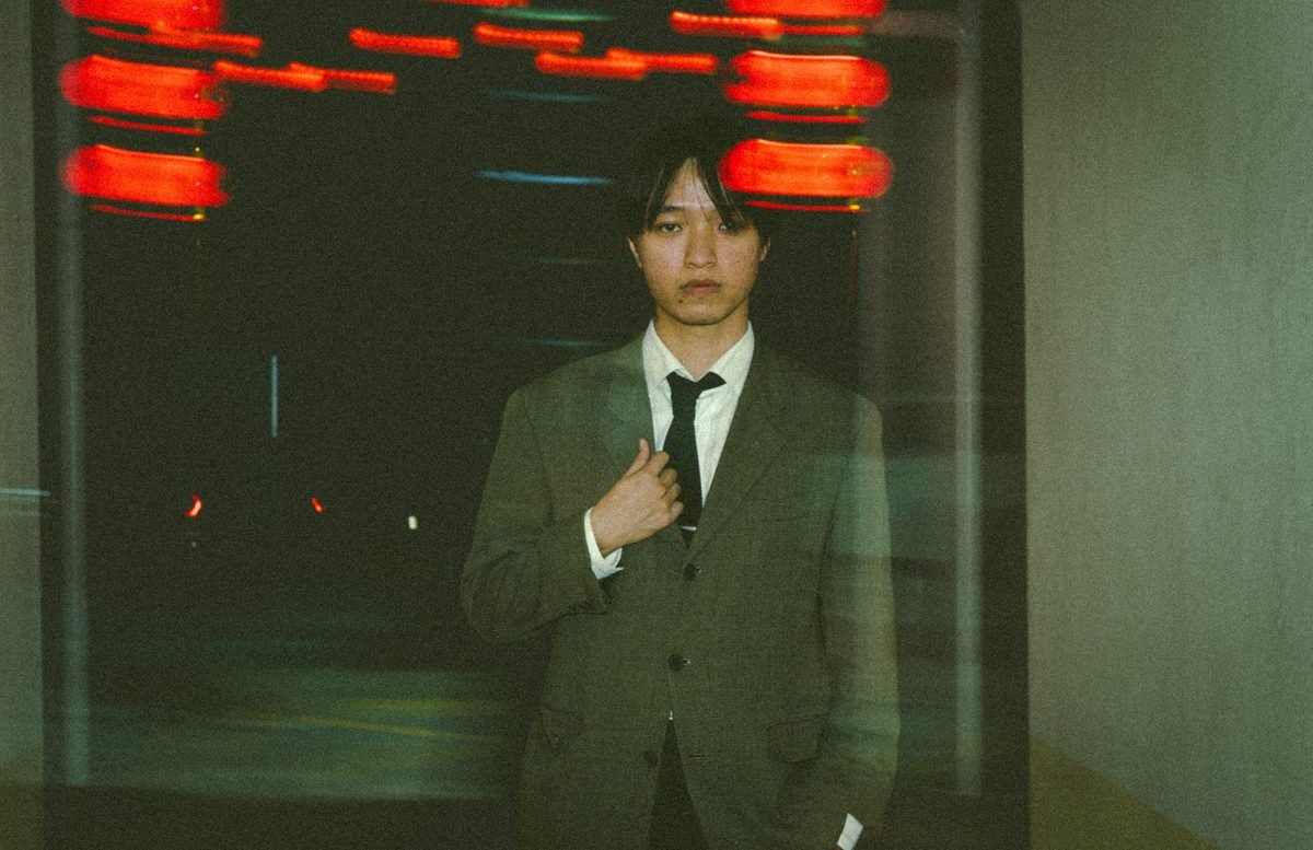 A man in a green suit jacket stands in front of a glass wall while red lights smear across the dark behind him.