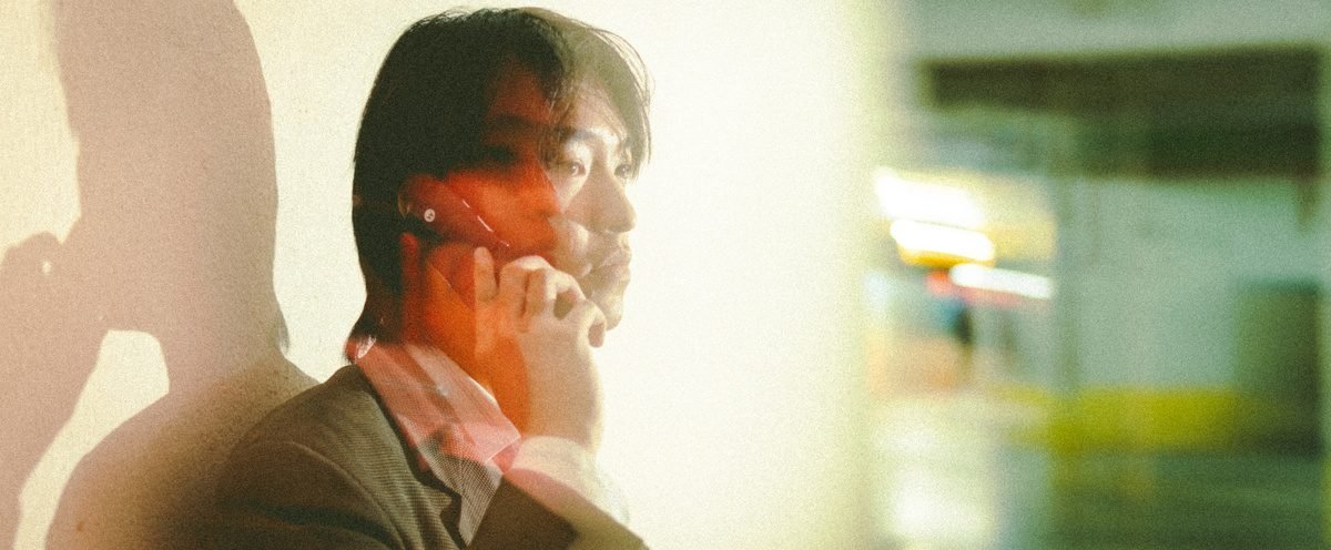 Close crop of a man leaning against a pale wall with a phone to his ear, orange light washing over his face.