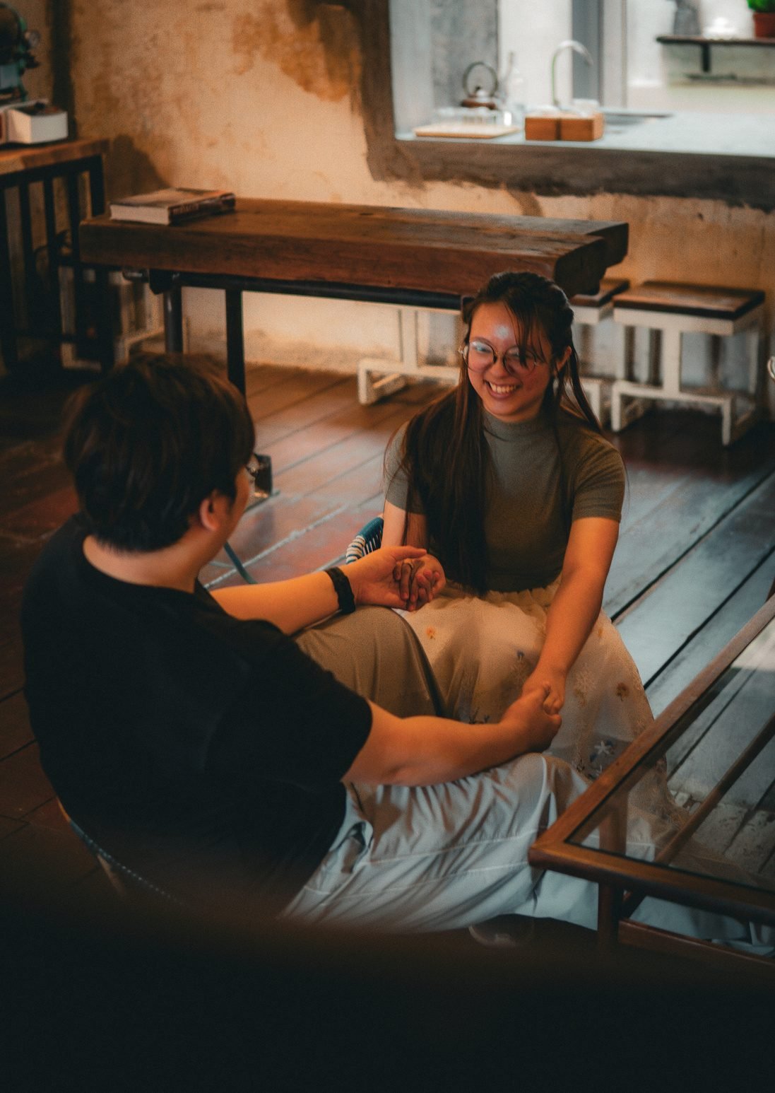 The same couple seen from above, sitting on a wooden floor in a cafe, holding hands.